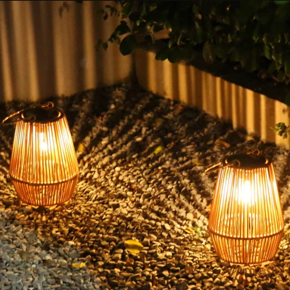 Two illuminated outdoor lanterns on a gravel path with a wooden fence in the background.
